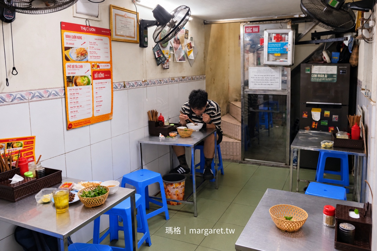 河內還劍湖附近 Tuệ An 蒸米皮捲店內環境,設有不鏽鋼餐桌、藍色矮凳與壁掛電風扇,牆上張貼蒸米皮捲、牛肉河粉及飲料菜單