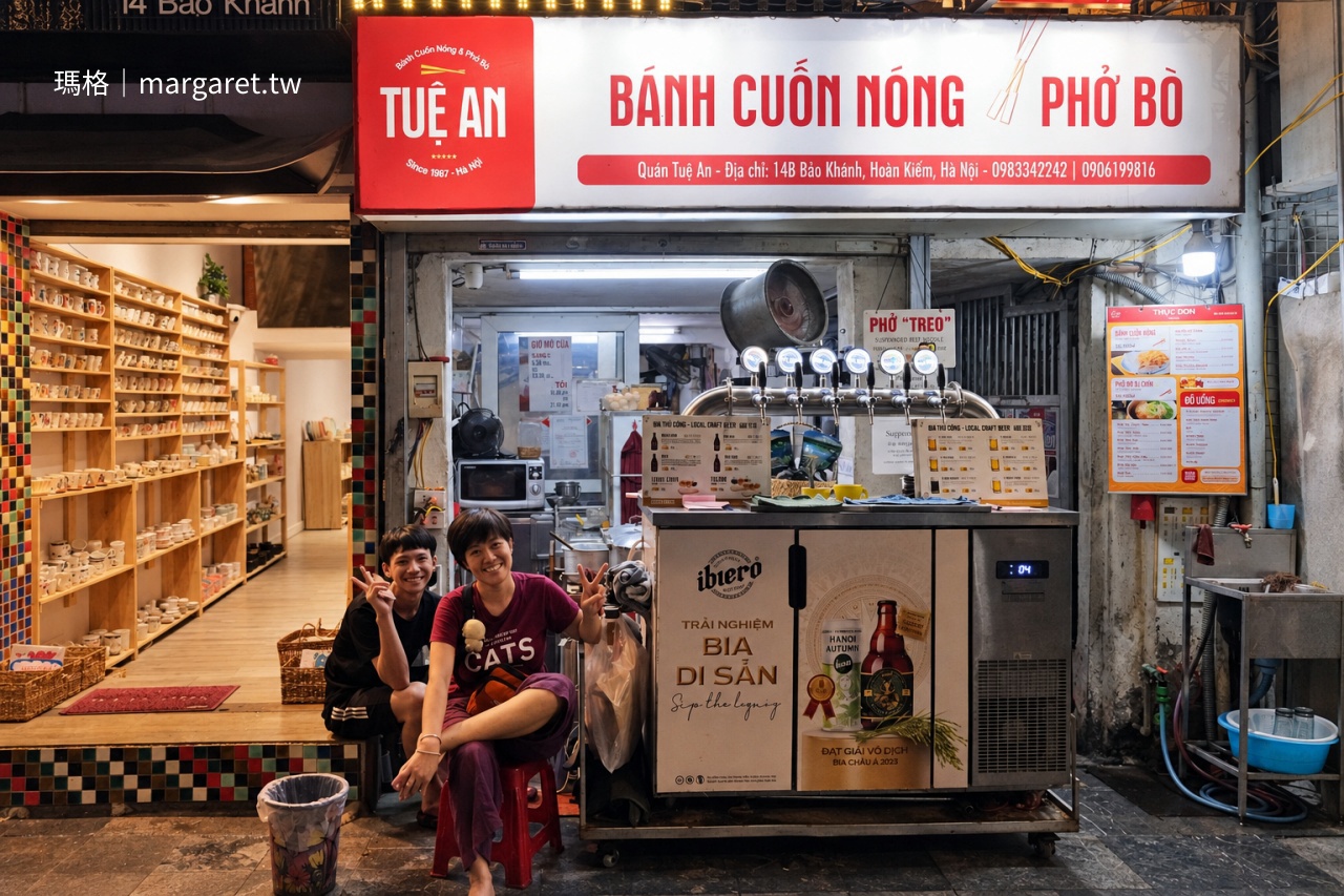 河內還劍湖附近 Tuệ An 蒸米皮捲店，老闆娘與兒子坐在店門口合照，身後是 Bánh Cuốn Nóng 與 Phở Bò 招牌及生啤酒吧台