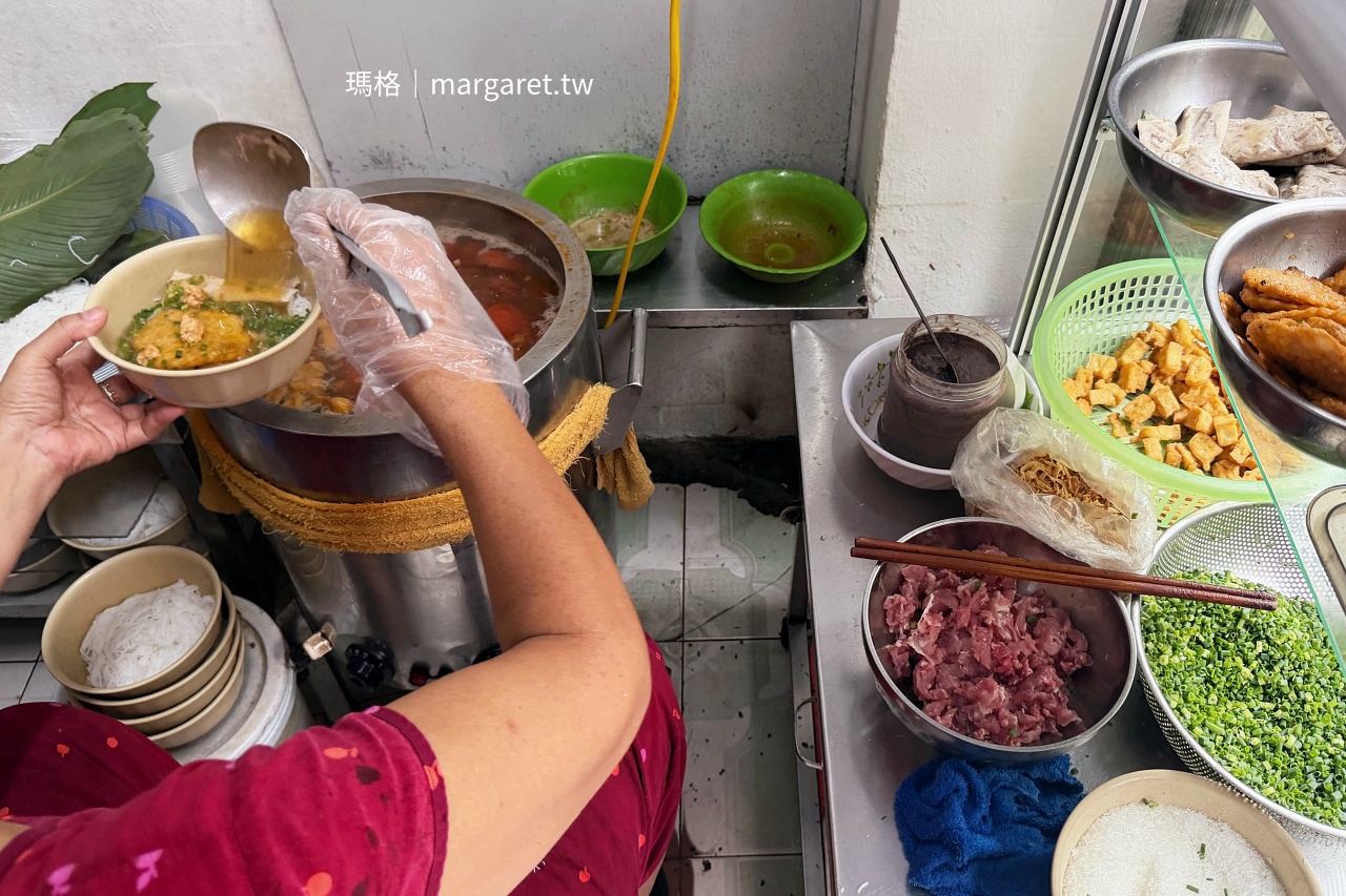 河內 Bún Riêu - Hàng Bạc 米線攤，老闆娘坐在門口大鍋旁舀湯，旁邊擺放食材櫃與調味料