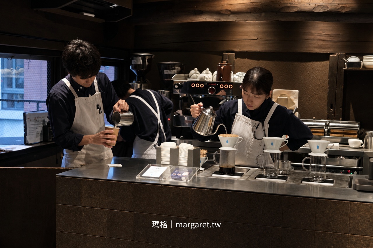 京都 Blue Bottle Coffee 六角店咖啡師專注製作飲品與手沖咖啡，以手沖壺向濾杯注水，吧台陳列白色咖啡器具