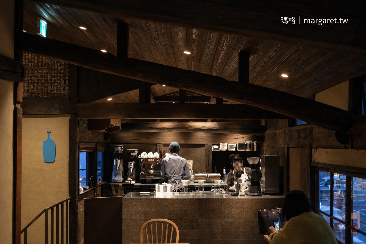 京都 Blue Bottle Coffee 六角店二樓俯瞰視角，老屋粗曠木樑下映照著暖光吧檯，咖啡師在吧台準備咖啡