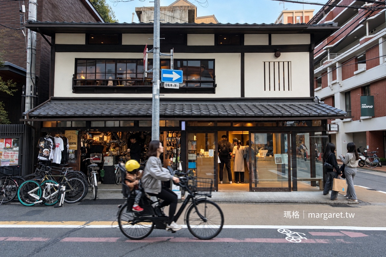 京都 Blue Bottle Coffee 六角店外觀，傳統日式町家白牆、黑色木構與瓦屋頂下，一樓為咖啡館與自行車店，行人及騎單車的親子經過街道
