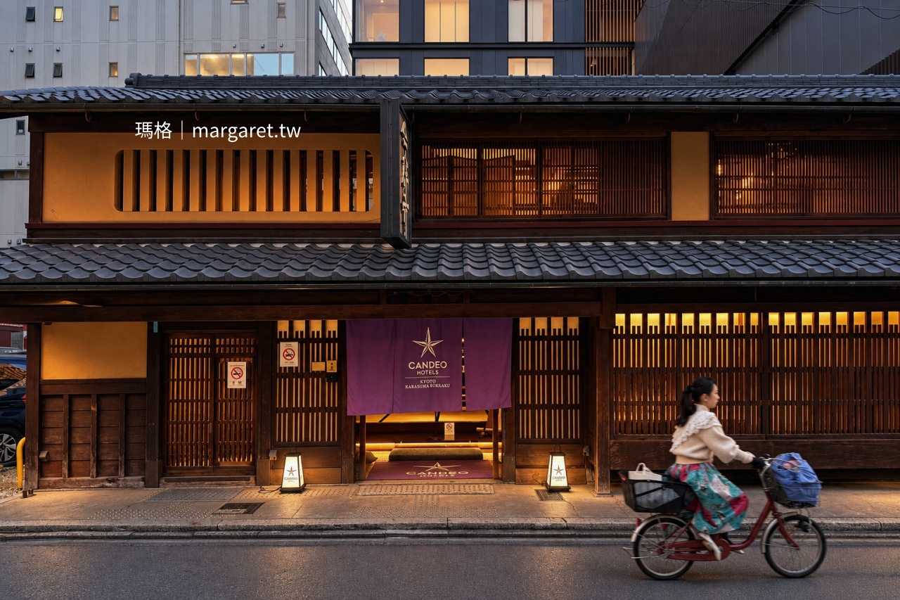 京都烏丸六角坎度京都烏丸六角光芒酒店（CANDEO HOTELS 京都烏丸六角）外觀，傳統町家木格窗、日式瓦屋頂與暖黃燈光，一名女子騎自行車經過酒店門前